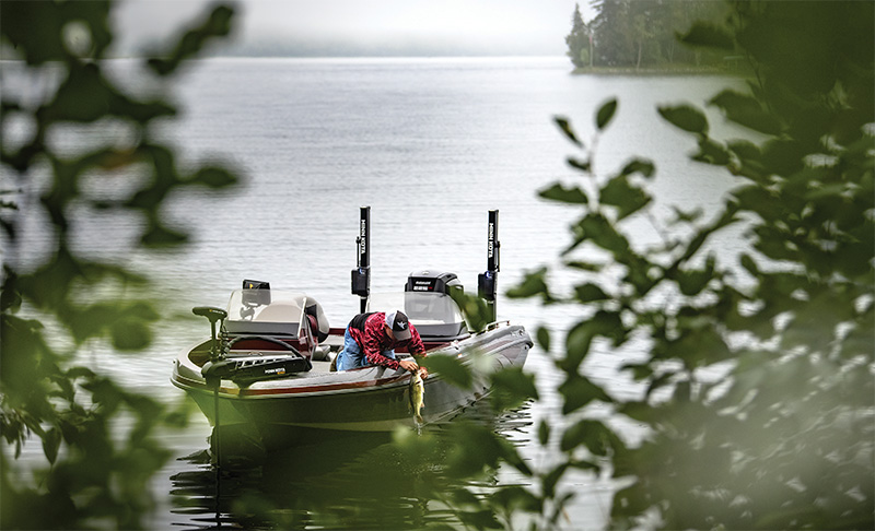 Angler auf einem Boot, umgeben von Bäumen, hält seinen Fang mit einem idyllischen See im Hintergrund.